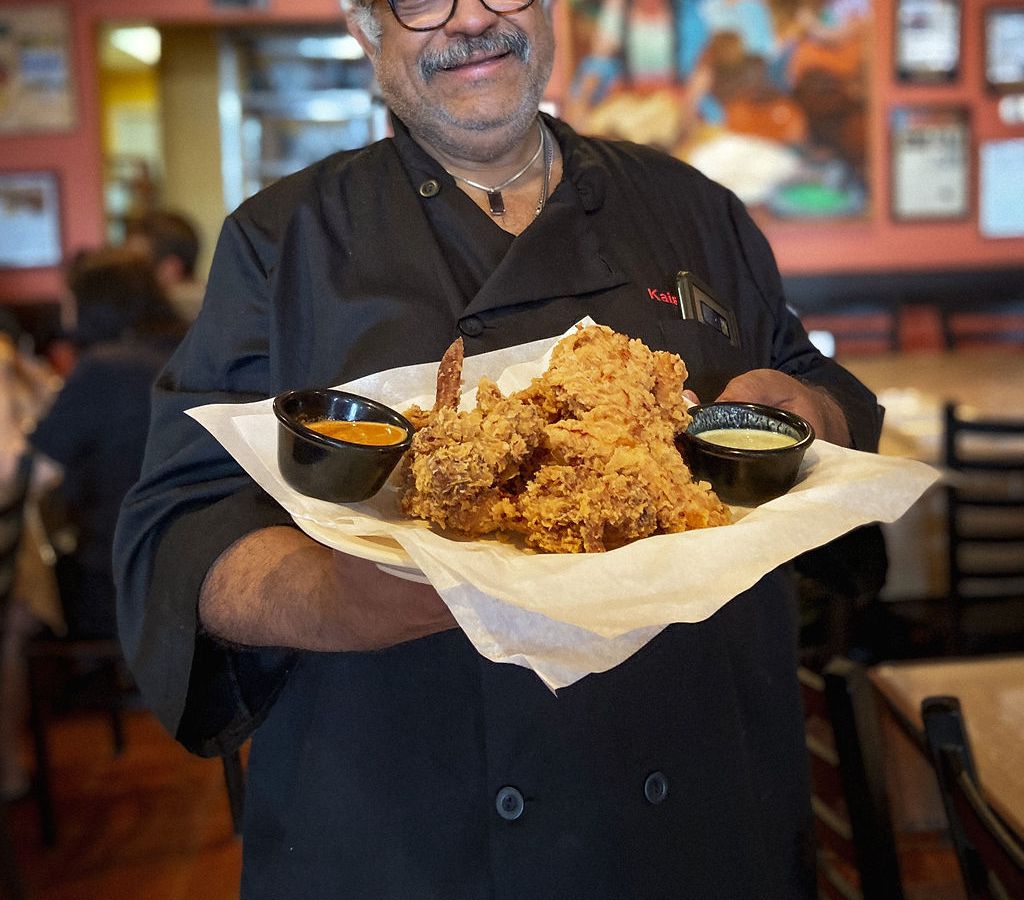 Wearing a black chef’s coat, Himalaya restaurant chef and owner Kaiser Lashkari holds a plate full of crispy-craggy fried chicken. In the background, a colorful mural of people at a celebration hangs on the wall surrounded by other framed portraits and certificates.
