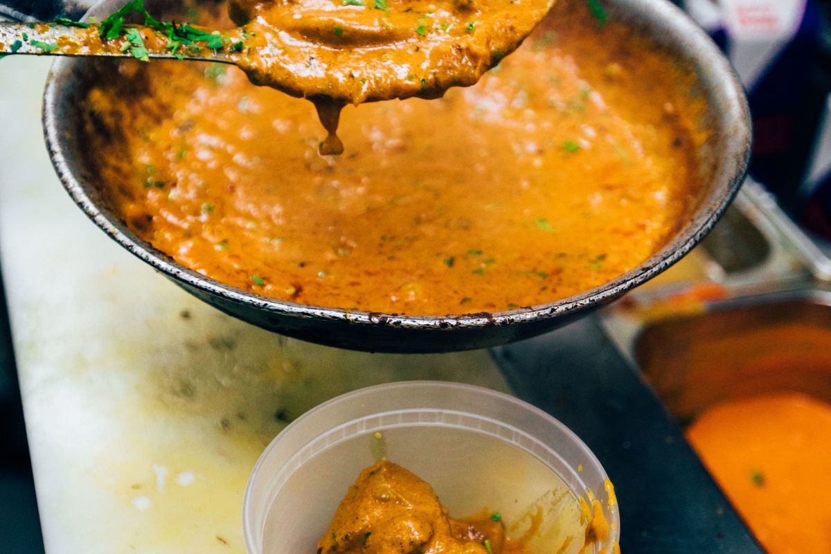 A metal kitchen spoon scoops bright orange butter chicken into a to-go container from a saucepan.
