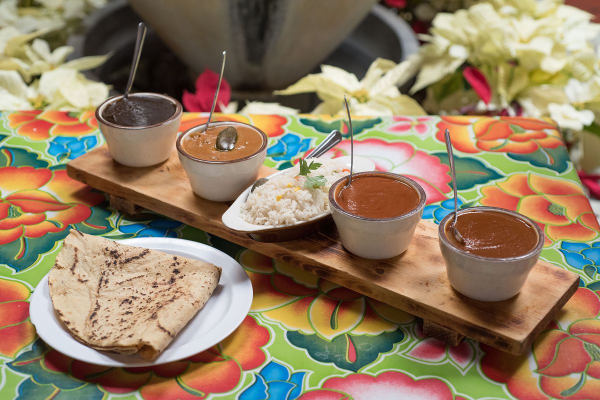 Four types of mole in small cups with a tortilla atop a colorful traditional tablecloth.