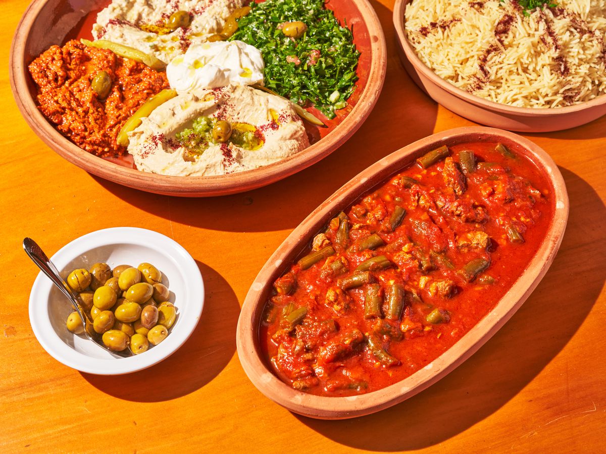 a red saucey plate of beans in a terracotta pot sits next to a colorful mezze platter on a tan table.