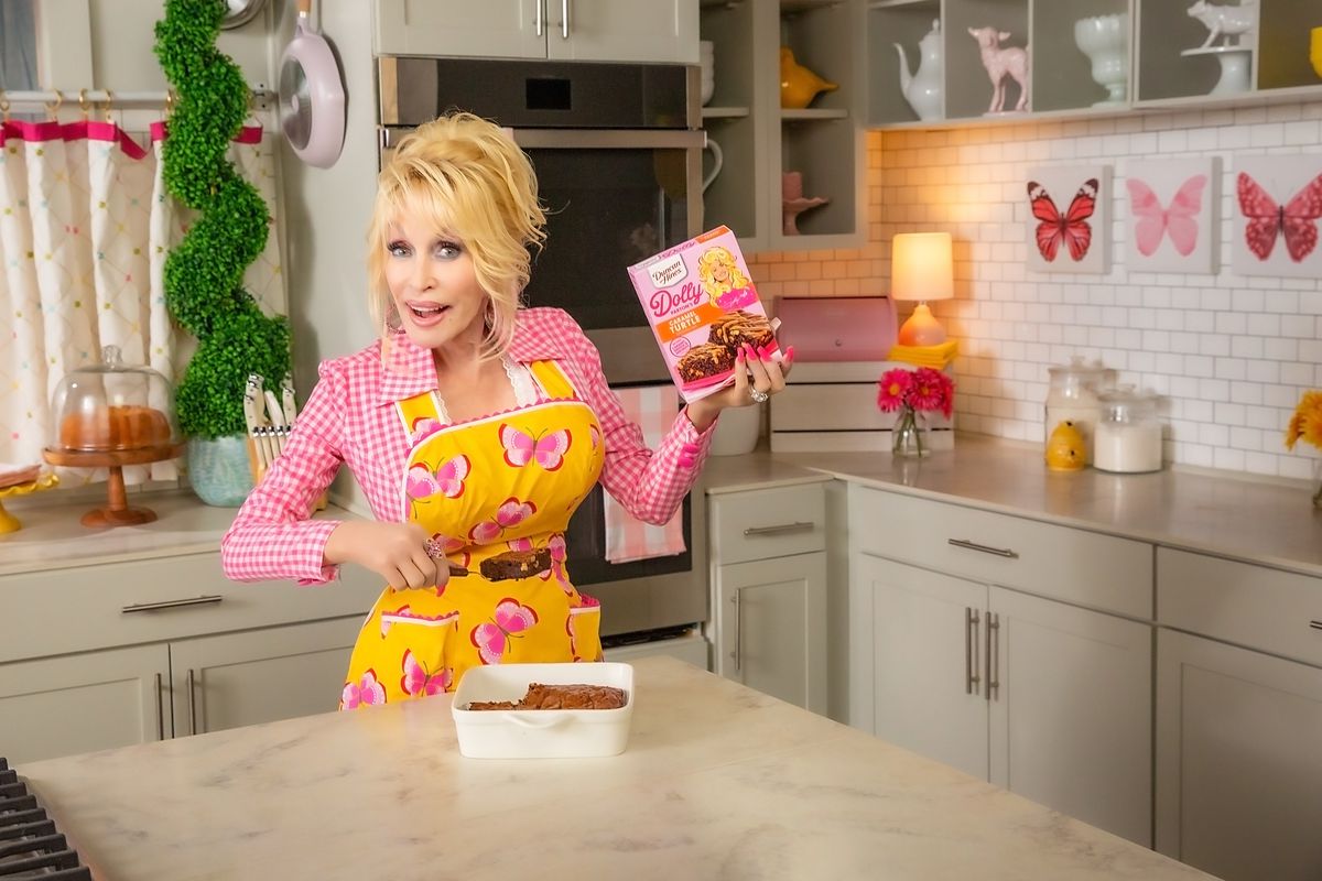 Dolly Parton, a blonde woman wearing a pink shirt and patterned yellow apron, stands in a kitchen holding pink boxes of cake mix. 