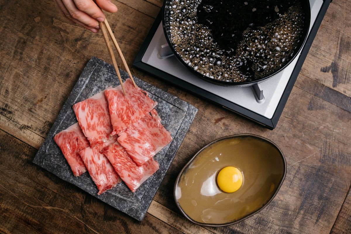 A person uses chopsticks to pick up raw pieces of wagyu to put in a boiling hotpot. Nearby, a raw egg sits in a bowl.