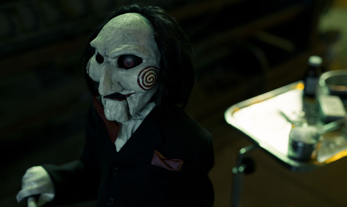 The signature Jigsaw puppet from the Saw movies, a red-eyed, white-skinned, black-haired humanoid with prominent cheekbones painted with red spirals, as seen in Saw X