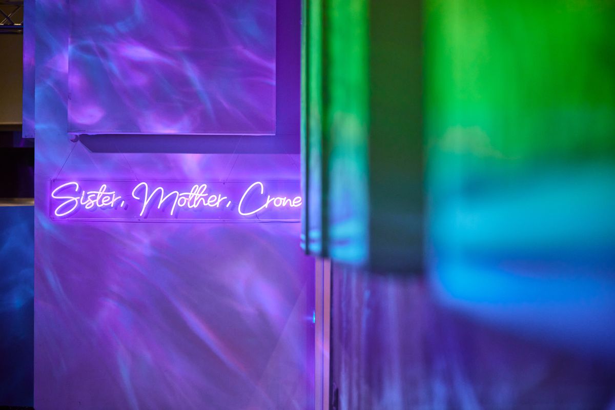 A neon sign reads “Sister, Mother, Crone” with prismatic art projections on the wall.