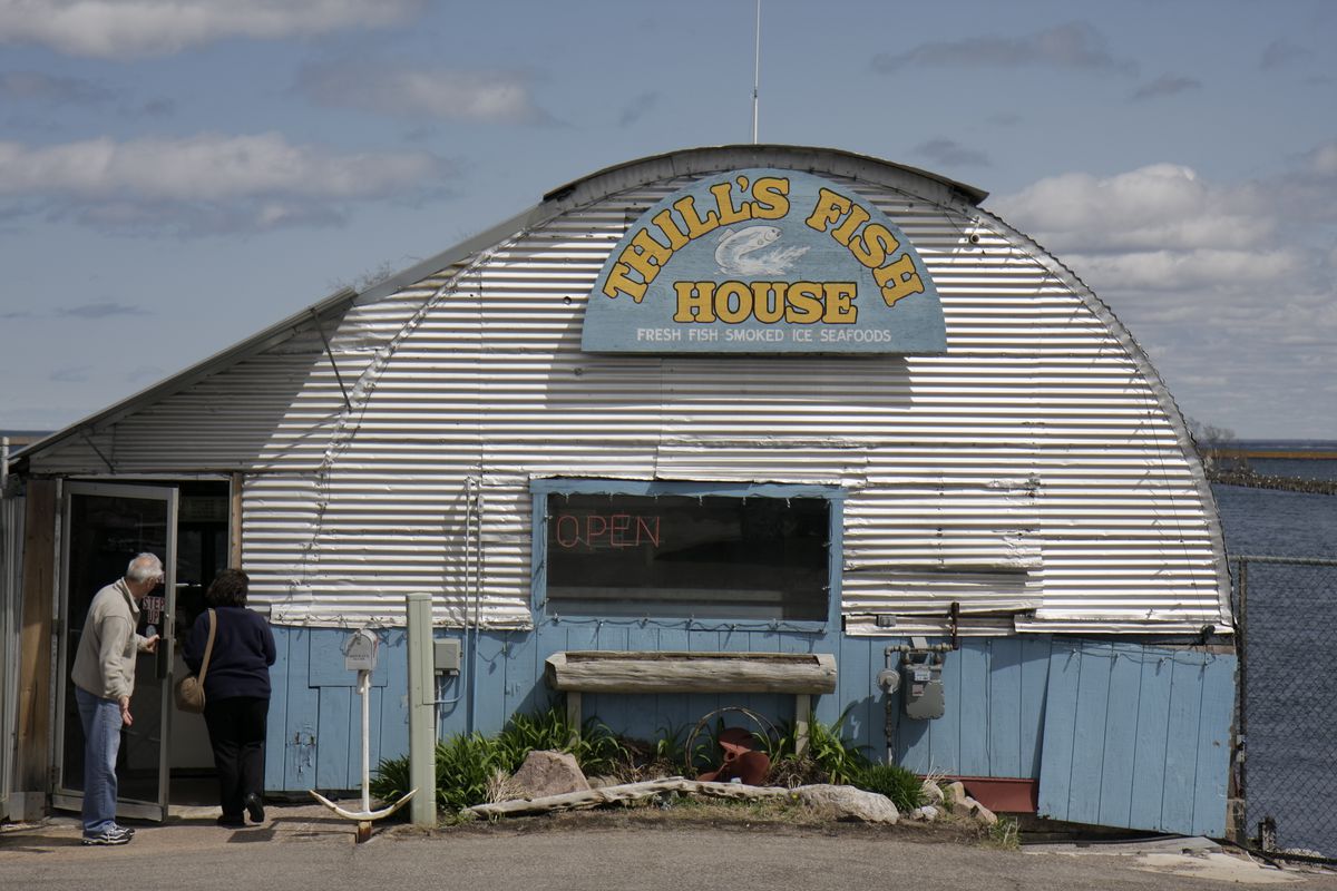 A customer holds the door for another entering a rounded metal building with slats of siding and a sign depicting a jumping fish that reads ‘Thill’s Fish House fresh fish smoked ice seafoods’