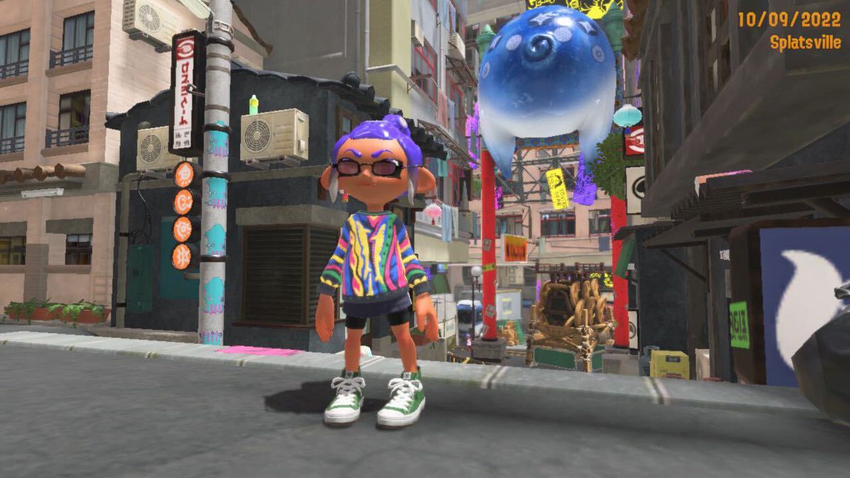 A purple-haired Octoling stands in the Splatsville city area of Splatoon 3. The Octoling is wearing a colorful sweater, stylish sunglasses, and green hi-tops.