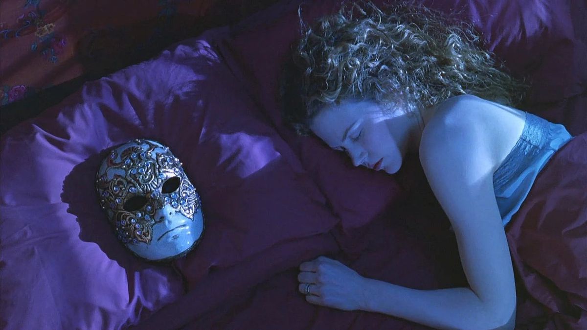 A woman (Nicole Kidman) sleeps in bed beside a jeweled masquerade mask placed on the pillow next her.