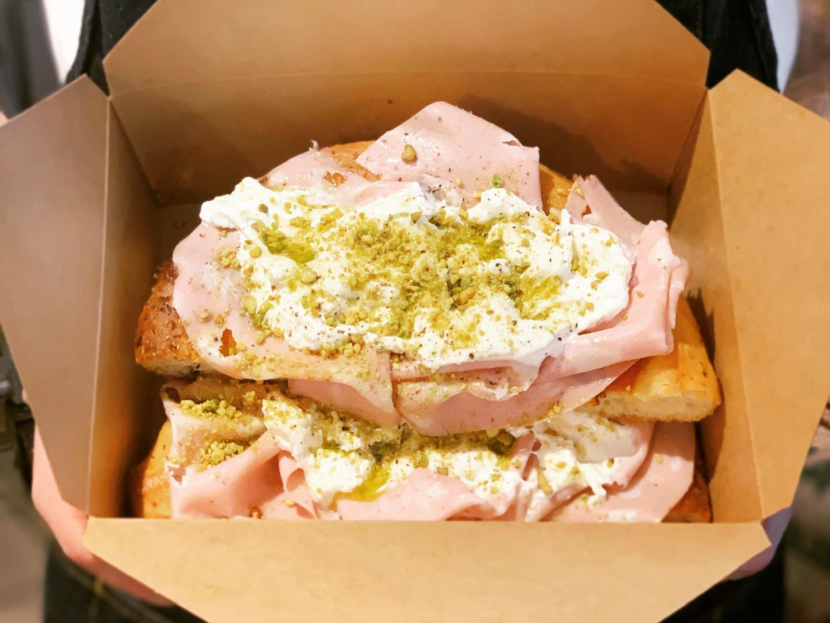 Focaccia topped with layers of mortadella, burrata, and chopped pistachios, presented in a cardboard takeout box.