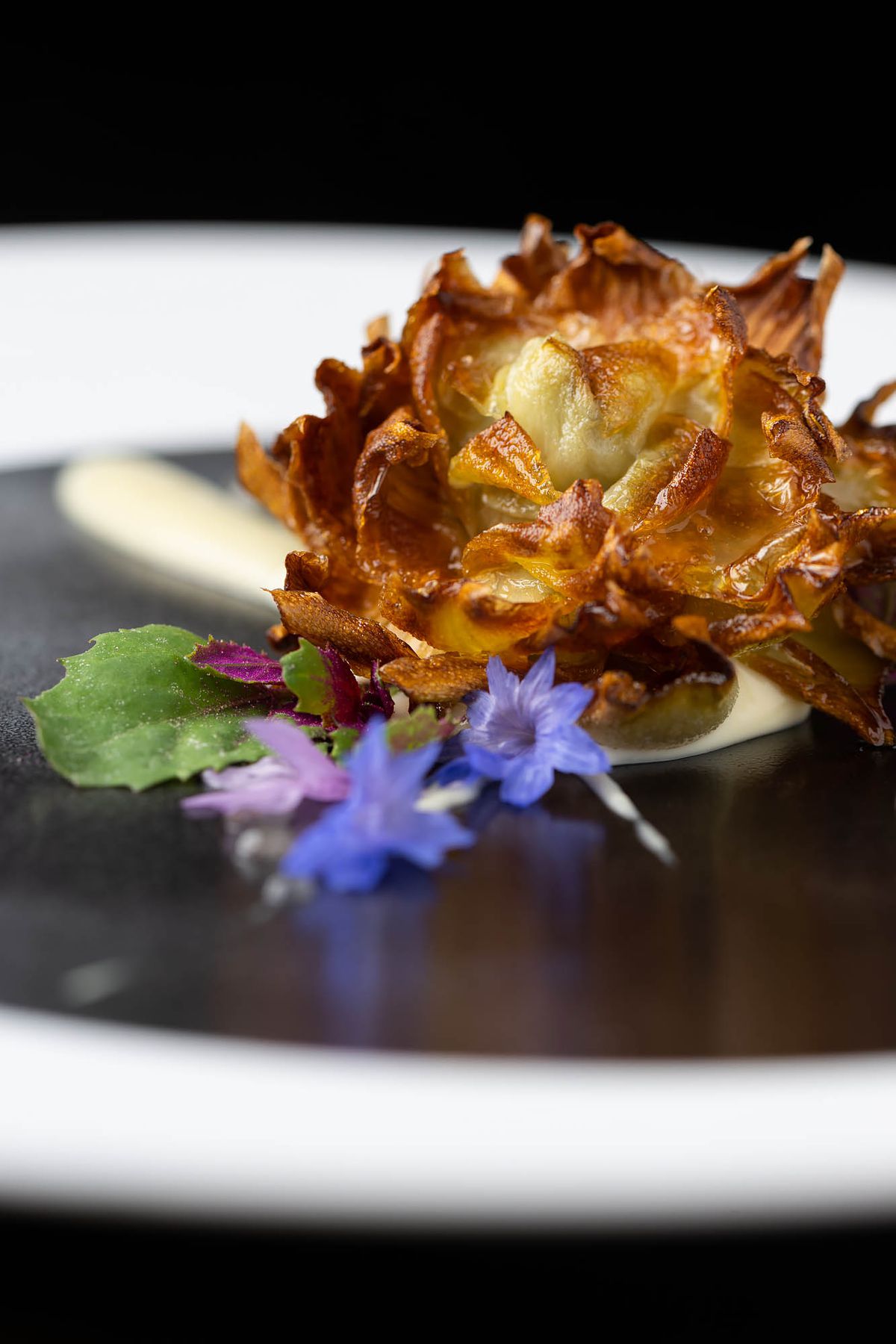 A black and white plate with a fried artichoke, miso, and Gruyere with little purple flowers at Pasjoli.&nbsp;