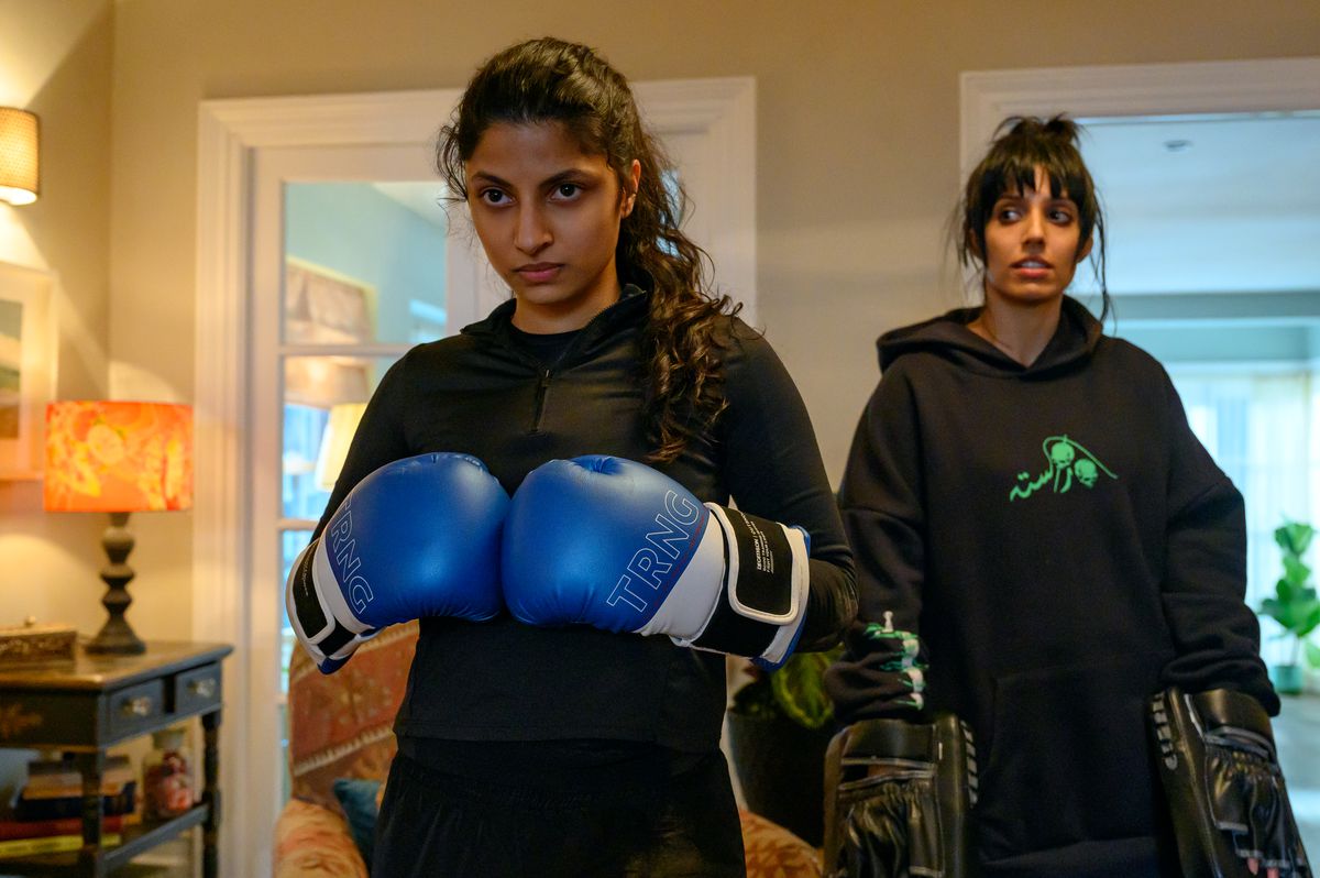 Ria, a British-Pakistani teenager, wears a black shirt and boxing gloves, which she presses against each other and looks determined. Behind her is her sister Lena, who wears a black hoodie and holds two strike pads for Ria to hit. They are currently down, though, as she regards Ria.