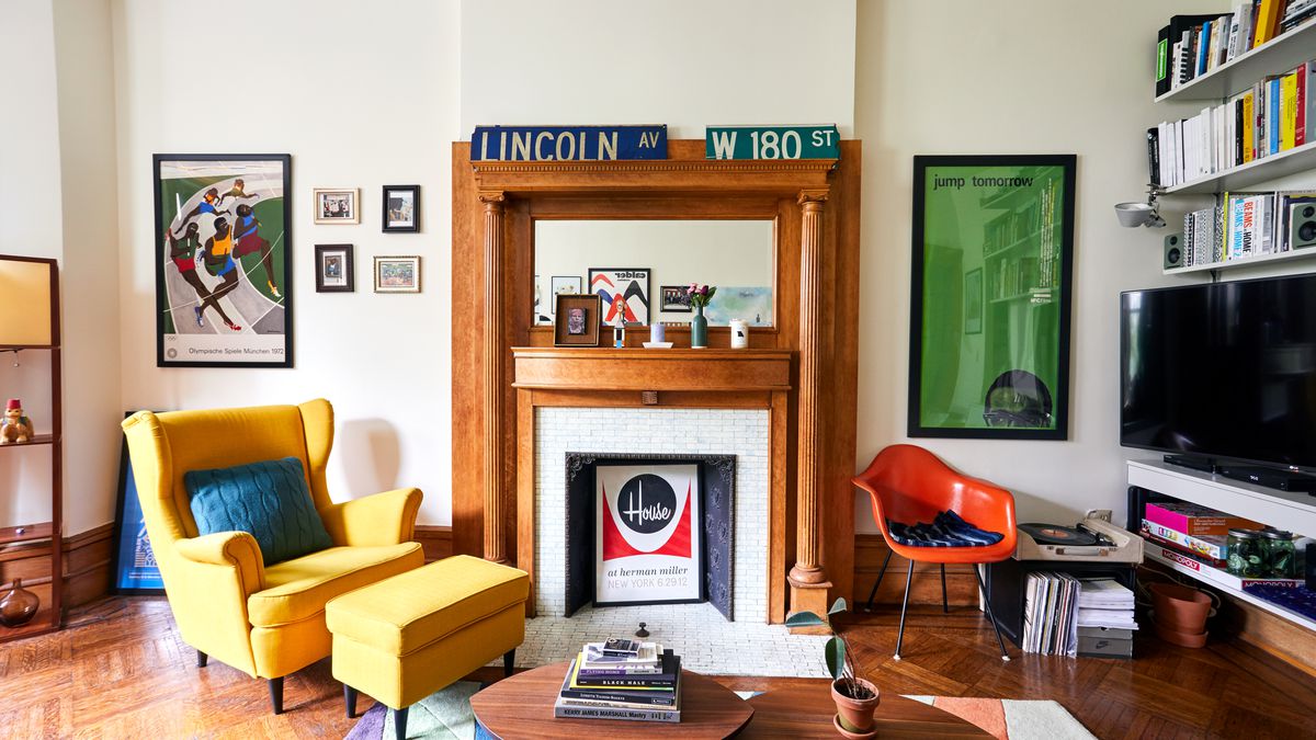 The living area of a New York City apartment with a fireplace at the center, a yellow lounge chair with footrest to the left, and a red accent chair and television to the right. Bold bright artworks hang around the walls.