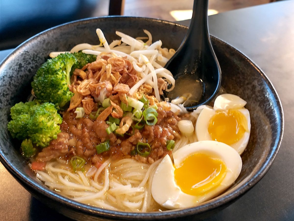 A bowl of noodles topped with boiled egg, broccoli, sprouts, and meat.