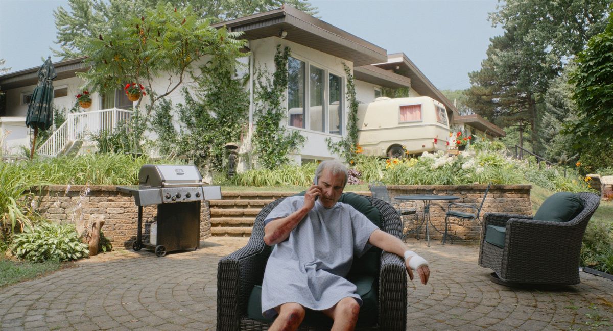 Joaquin Phoenix, in a hospital gown, sitting on a patio chair, with an ordinary suburban house in the background.