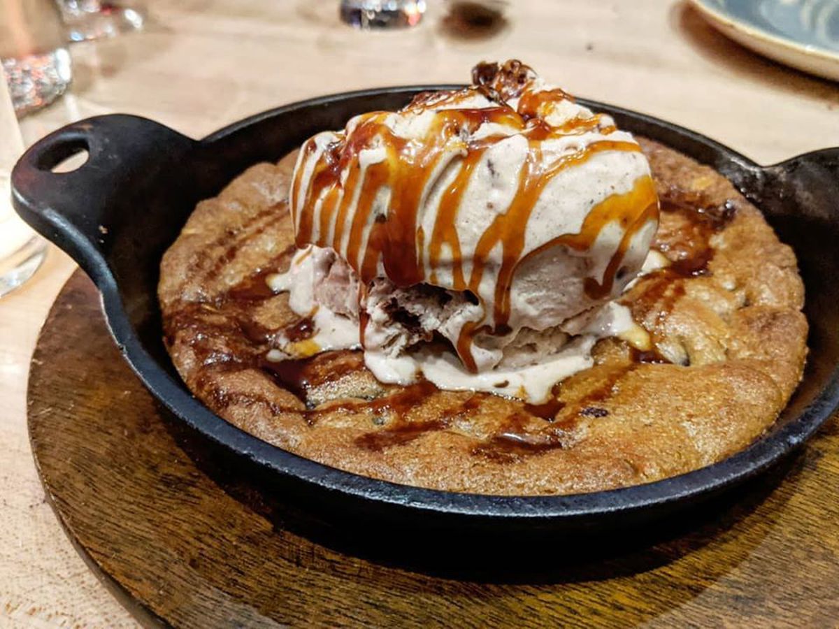 A cookie baked in a cast-iron skillet is topped with a scoop of ice cream and a drizzle of caramel