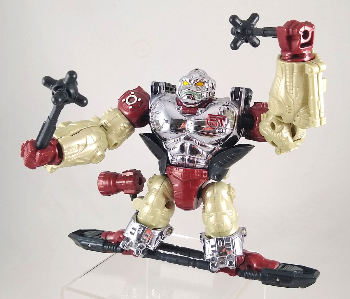 A photo of an Apelinq convention exclusive Transformers toy, a red, black, silver, and cream metal ape standing on a black hoverboard and wielding black maces