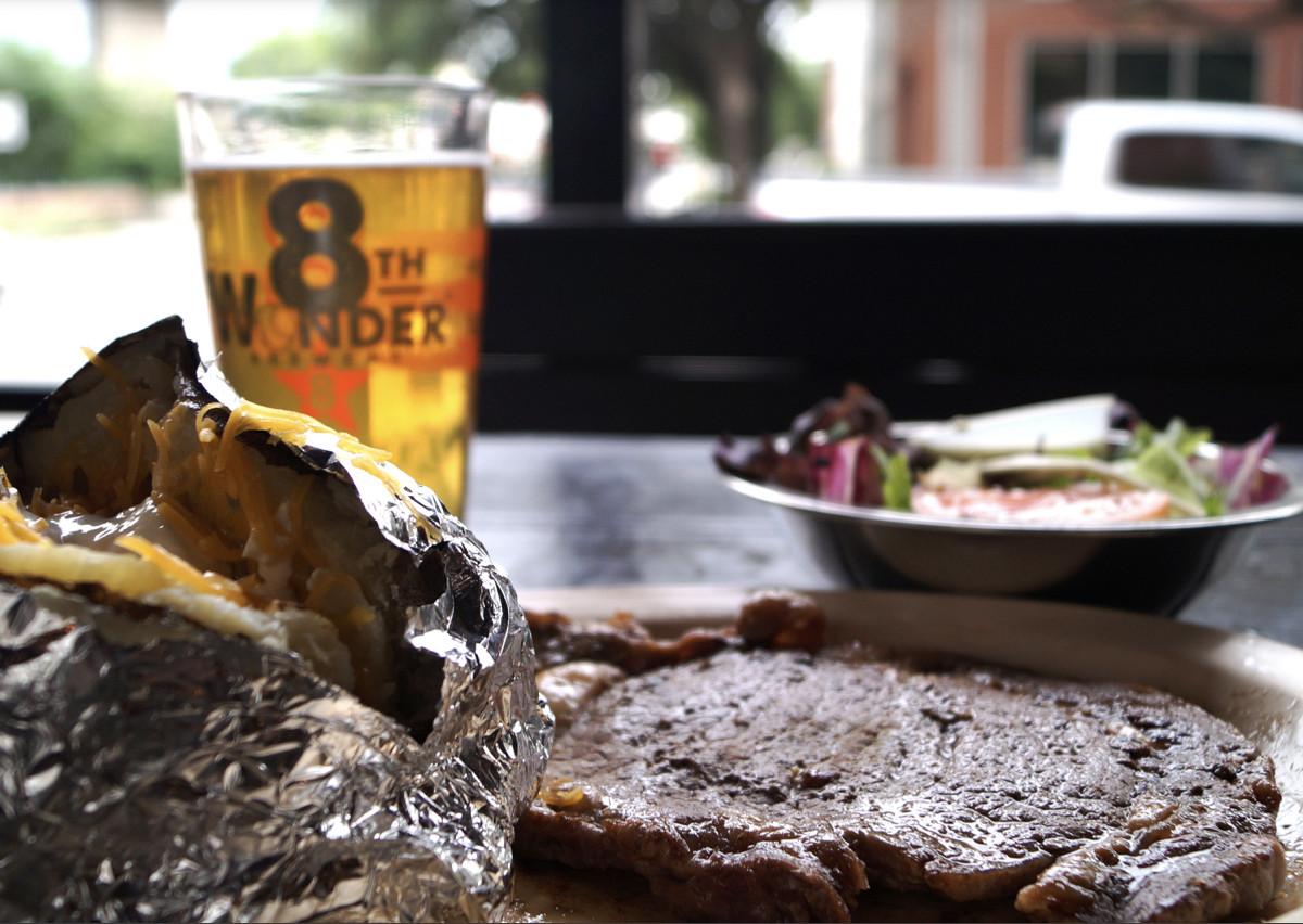A steak served with a foil-wrapped baked potato, side salad, and pint of 8th Wonder Brewery beer.