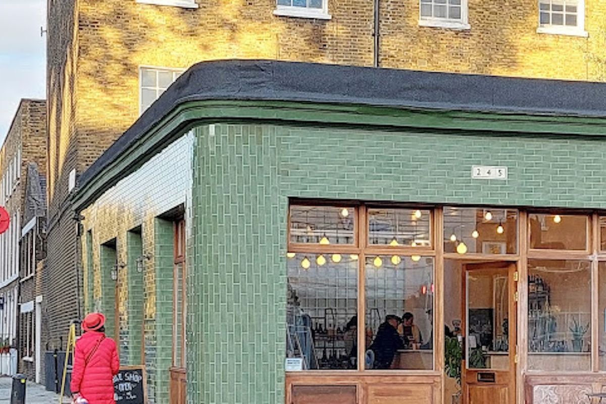 A green-tiled corner unit with wooden framed windows, and a gently curved exterior. A chalkboard sits outside, with a woman in a red coat walking and admiring the frontage.