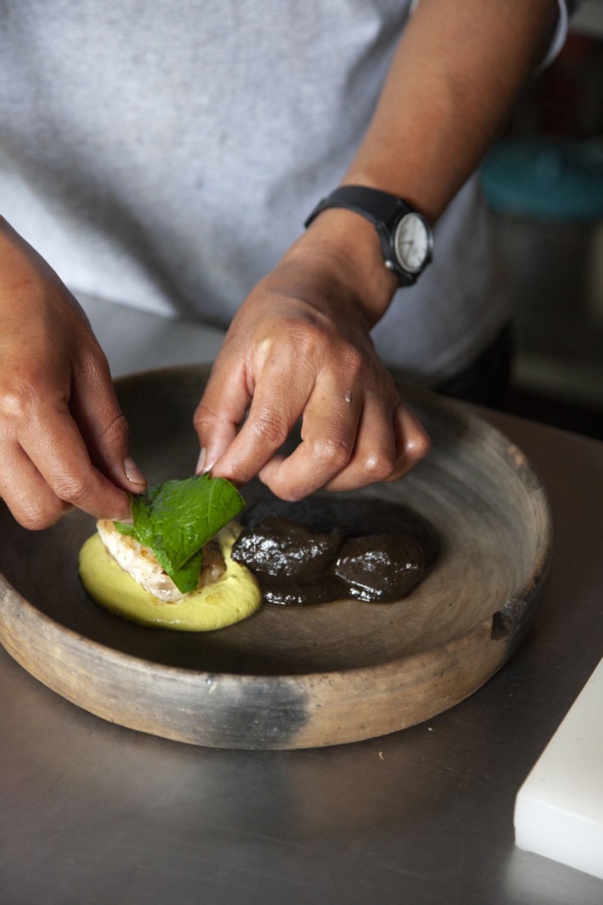 One bright green mole on a plate alongside a dark brown mole. Two hands place a piece of fish and leaf on top.