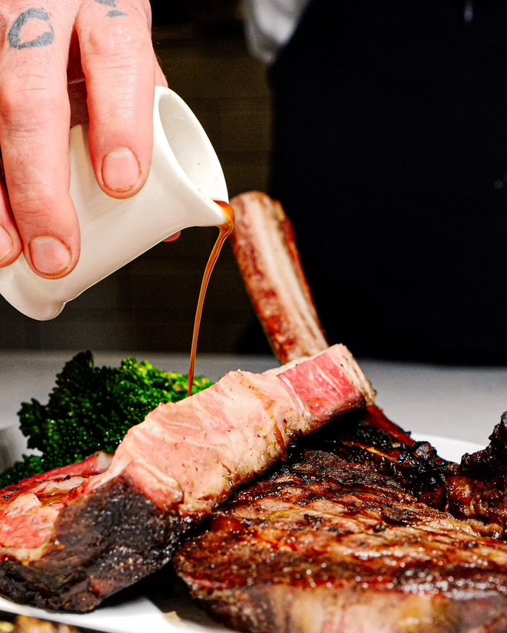 A hand pours brown sauce from a pitcher onto a plate of medium-rare beef