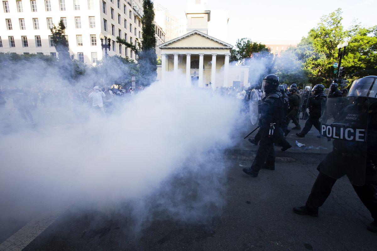 Buildings and protesters are obscured by a dense cloud of white gas — the only clear area, to the right of the photo, shows police in riot gear marching forward.