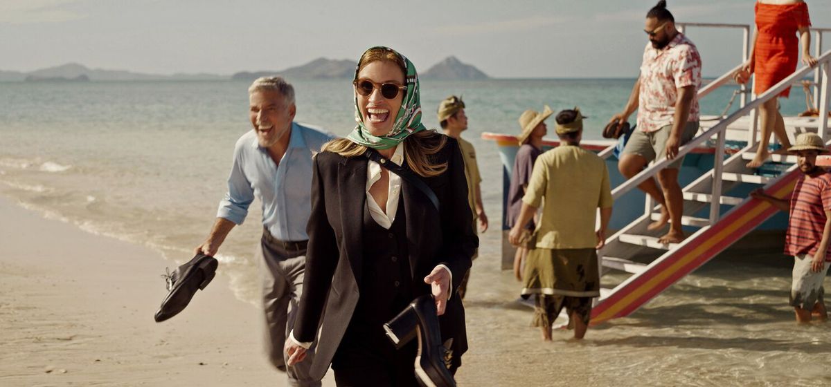 A man (George Clooney) and a woman wearing a scarf over her head (Julia Roberts) laugh while disembarking a boat onto a beach and holding their shoes in their hands.