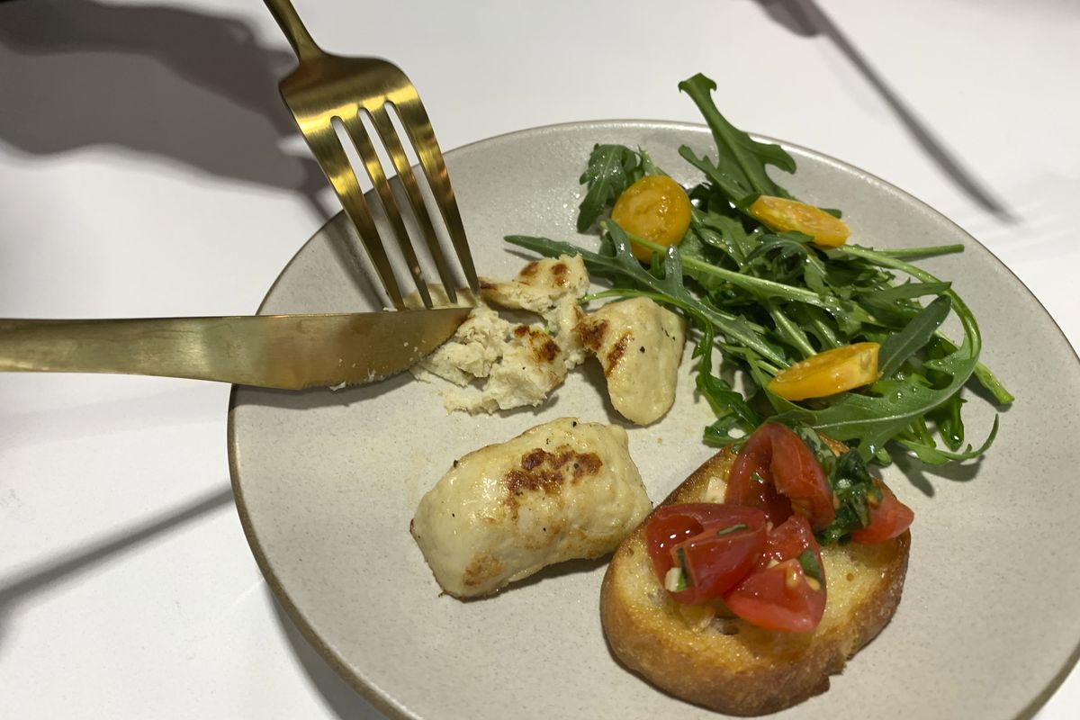 A knife and fork cutting a piece of lab-grown chicken meat on a dinner plate with greens and bruschetta.