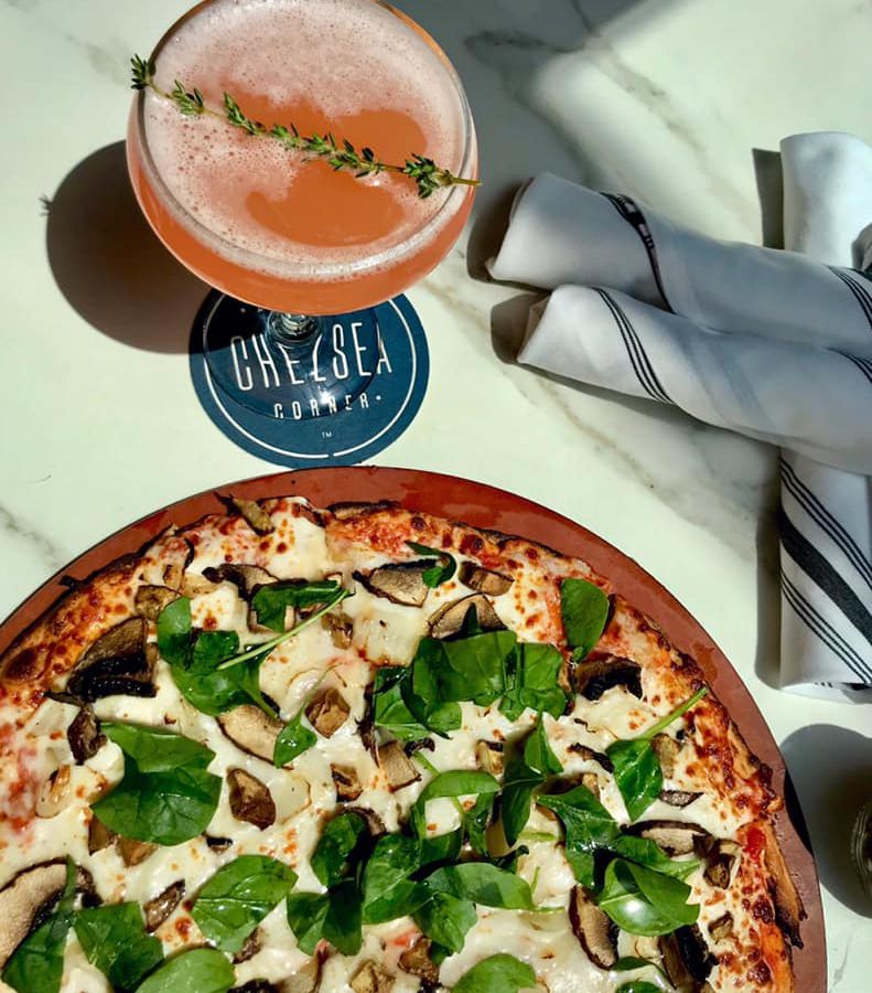 Gluten-free pizza topped with spinach and mushrooms, pink cocktail in the background