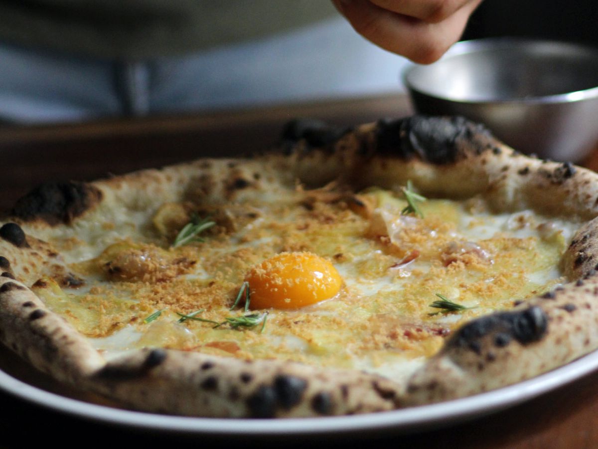 A chef adds herb fixings to a pizza, which features a bold yellow egg yolk in the center.