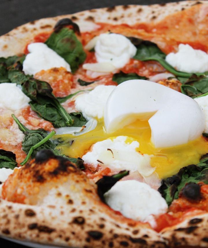 A poached egg cut open so the yellow yolk runs out onto a charred cheese pizza with greens on top.