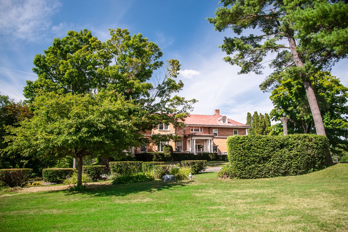 Large light-brick mansion with columned portico set behind shrubs, trees, and a lawn. 