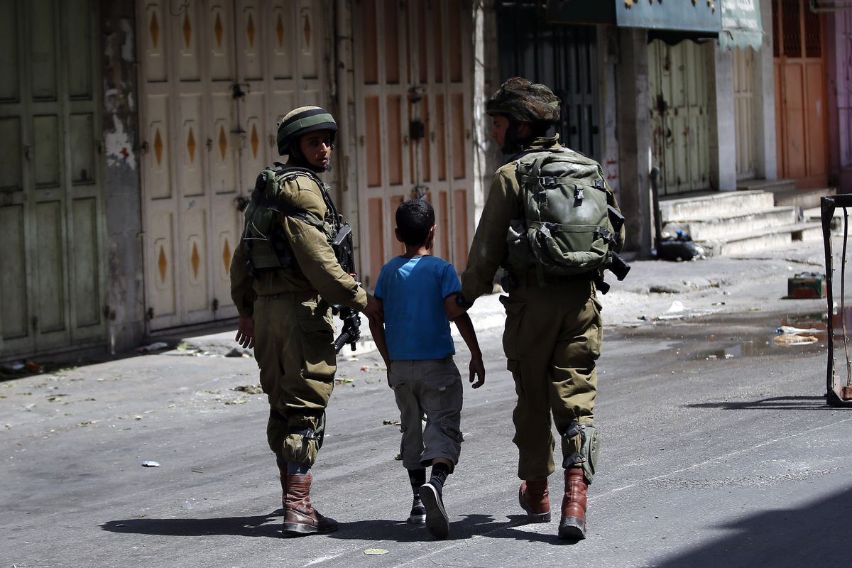 Two Israeli soldiers are photographed from behind as they hold a young Palestinian boy by his arms before detaining him.