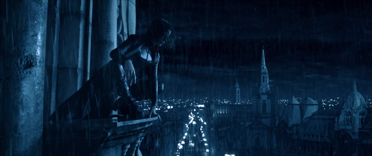 A short-haired woman in a leather jumpsuit and black coat kneels on the edge of a building overlooking a city at night with church steeples and buildings in the distance.