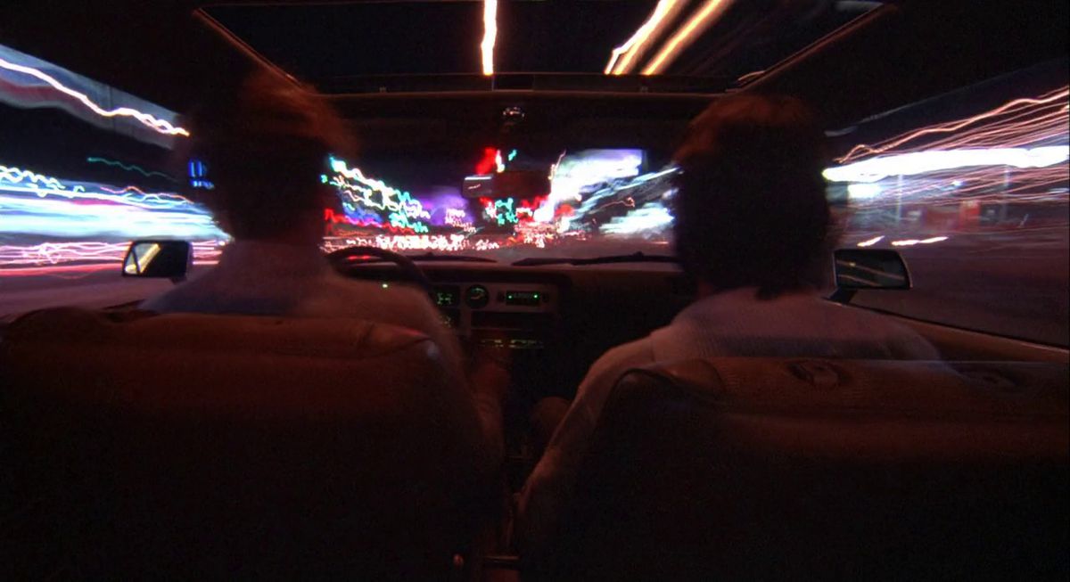 A man and a passenger behind the wheel of an automobile as streaks of light trail around them in Koyaanisqatsi.