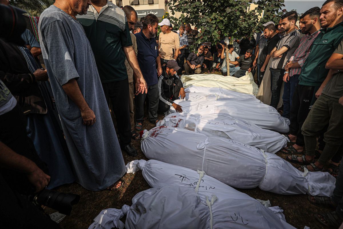 Bodies in white bags lie on the ground while a crowd of men looks on.