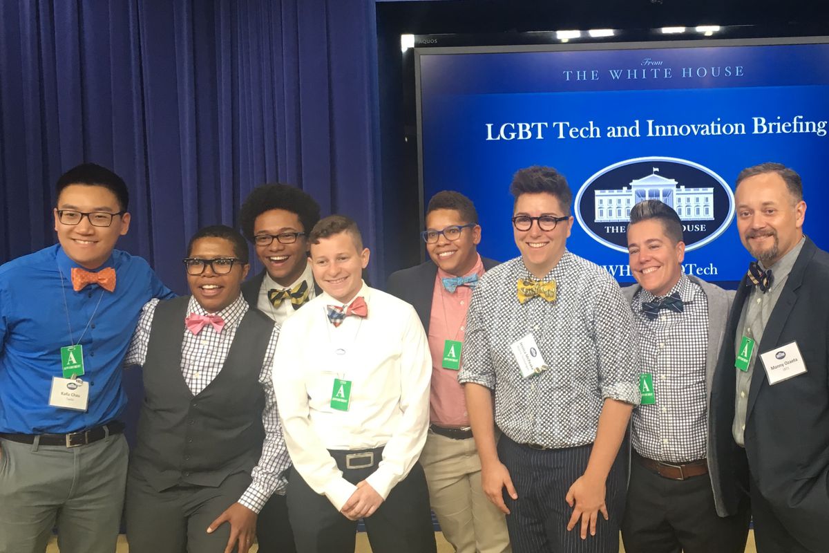 Eight people, a mix of races and genders, grinning and wearing casual shirts with bow ties in front of a White House backdrop. 