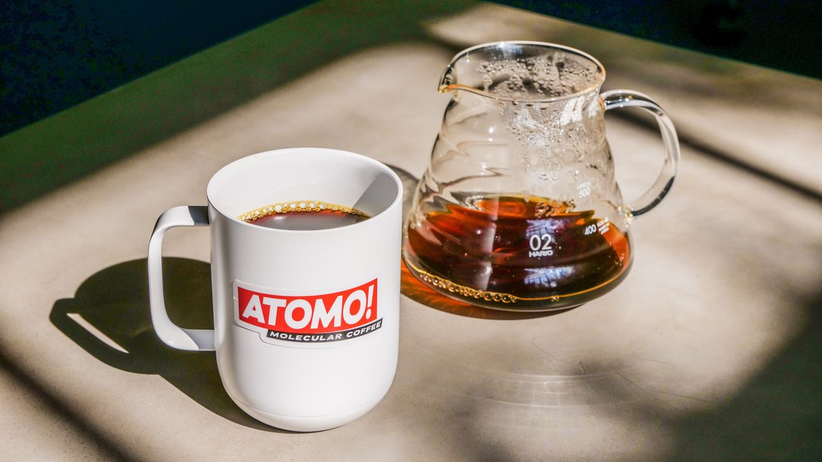 A white mug with the Atomo logo in red next to a glass beaker filled partly with coffee.