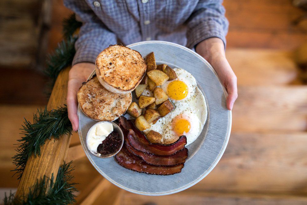In an overhead shot, two hands hold a light-colored plate filled with bacon, eggs, homefries, and English muffins.