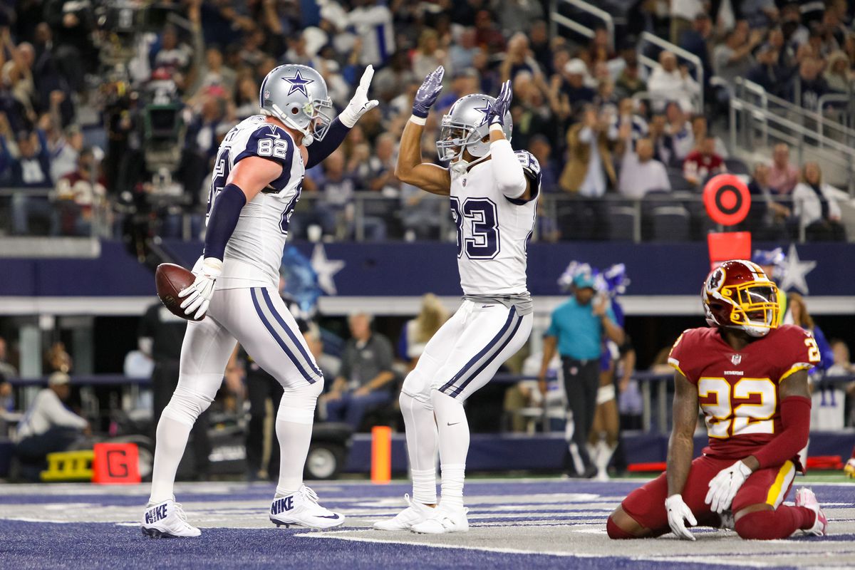 Two Dallas Cowboys football players high-five one another in the end zone after a touchdown while a Washington Redskins player slumps in defeat.