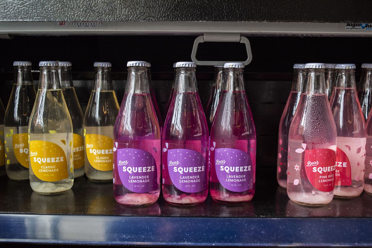 Pink, purple, and yellow beverages bottled in clear glass set in a refrigerated display case.