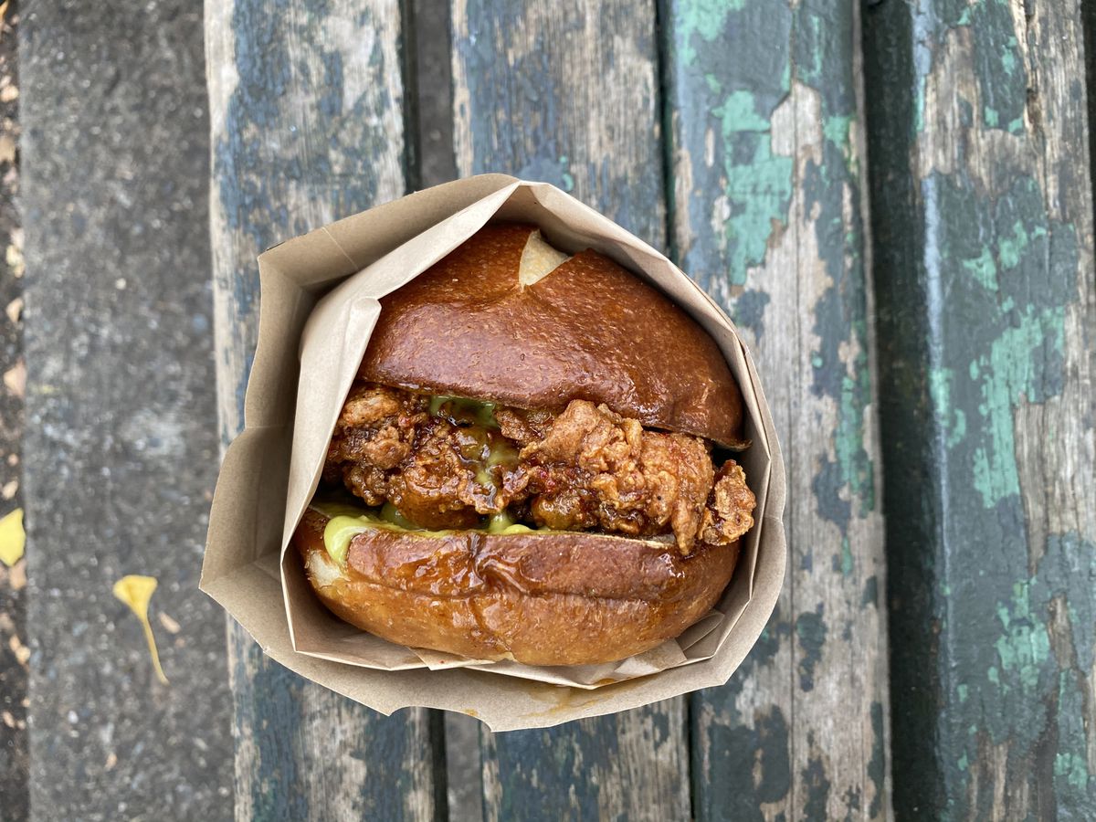 A fried chicken sandwich on a pretzel roll is shot from overhead in its packaging, with green Thai basil sauce leaking out from underneath