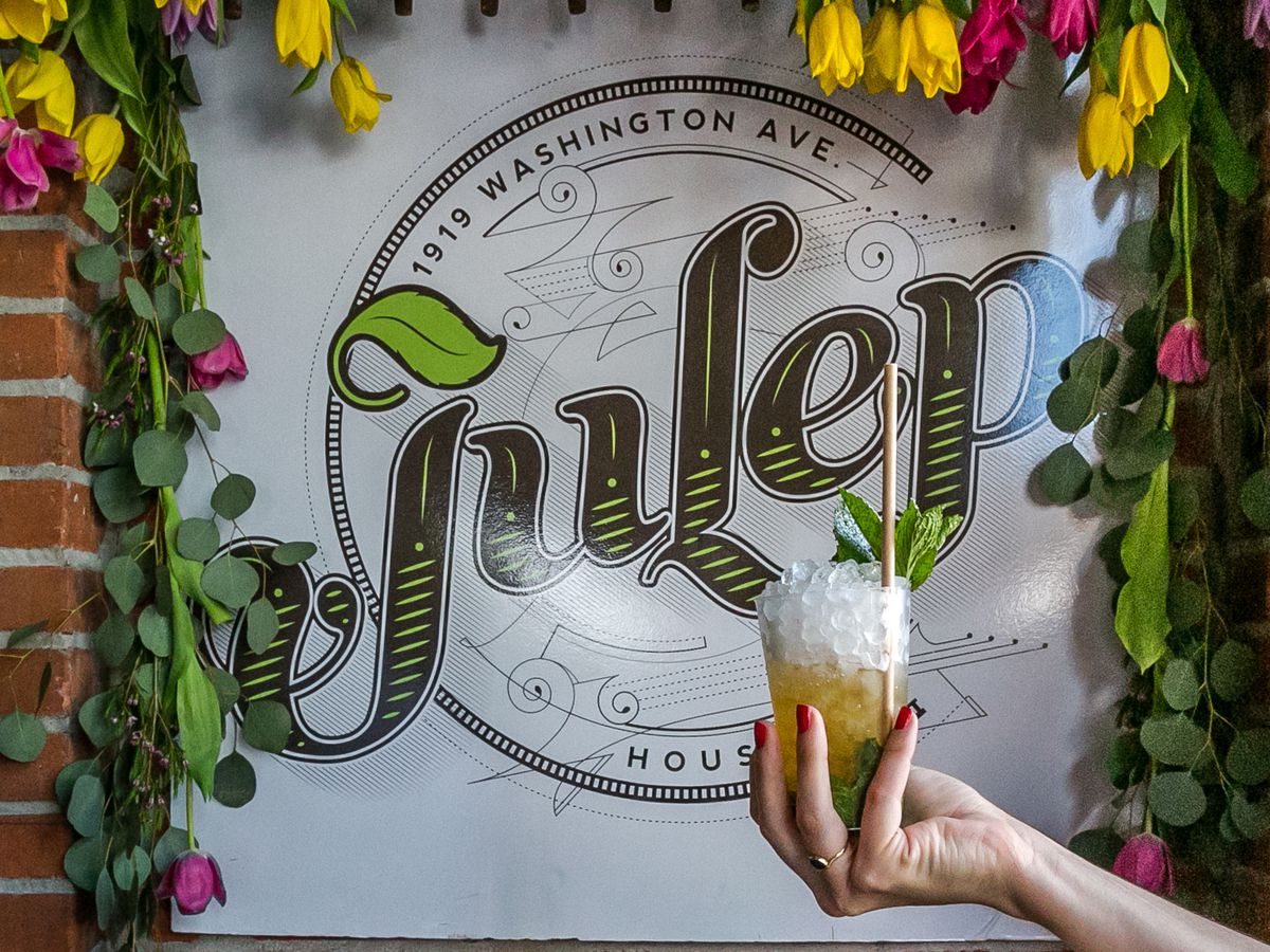 A hand holds a cocktail garnished with mint leaves in front of the Julep sign, which is framed in flowers and greenery.