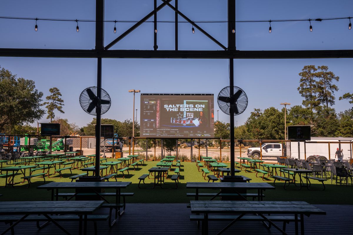 An inside view of Kirby Ice House - the Woodlands’ outdoor patio, which features a 300-inch jumbo screen, multiple picnic tables, and fans.