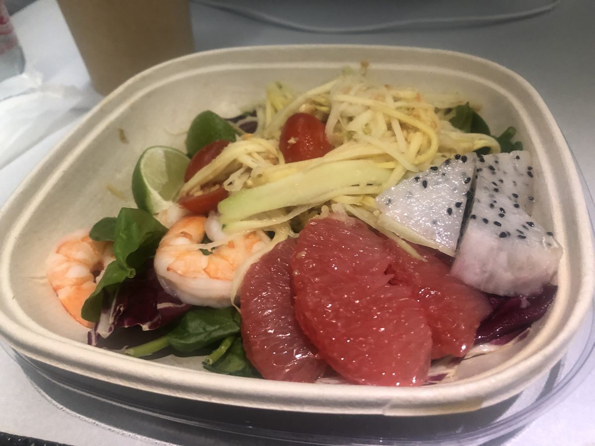 A takeout container filled with shrimp, mango, papaya, cherry tomatoes, grapefruit, dragon fruit, and two lime wedges.