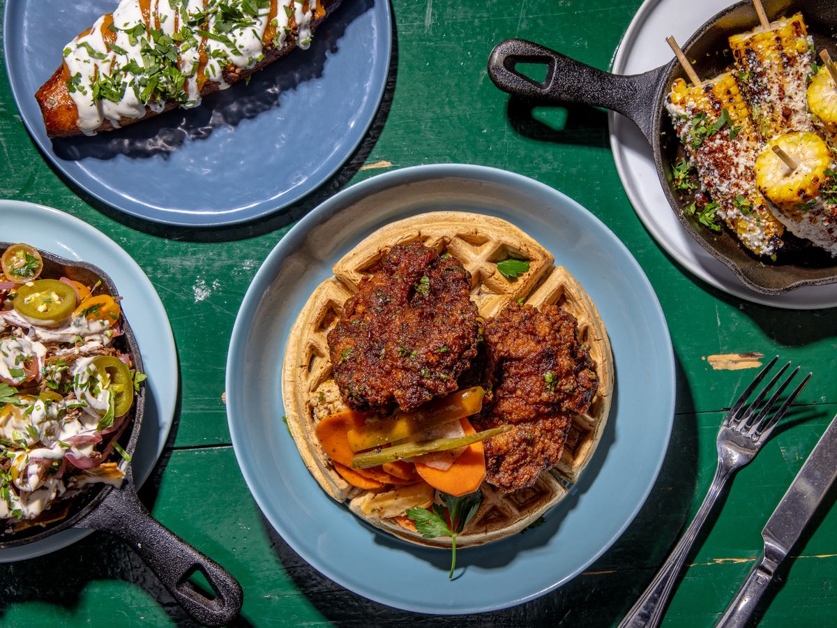 Golden sumac chicken over falafel waffles sits in the center of the photograph in an overhead shot; a variety of other dishes flank the chicken, including maduro mahshi, plantain nachos, and Mexican corn