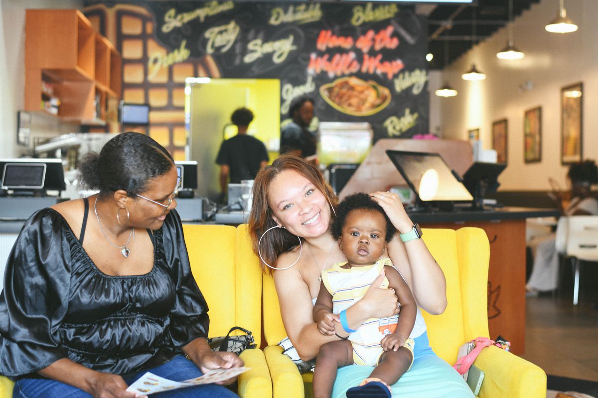Two women, one wearing a black shirt and looking at a baby seating in the other woman’s lap, the other woman wearing blue smiling. Both seated on yellow upholstered seating at the Waffle Cafe in Detroit, Michigan.