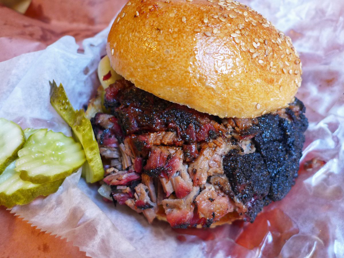 Black and pink rimmed beef brisket piled high on a sesame seed dotted roll, with green pickles tumbled on the side.