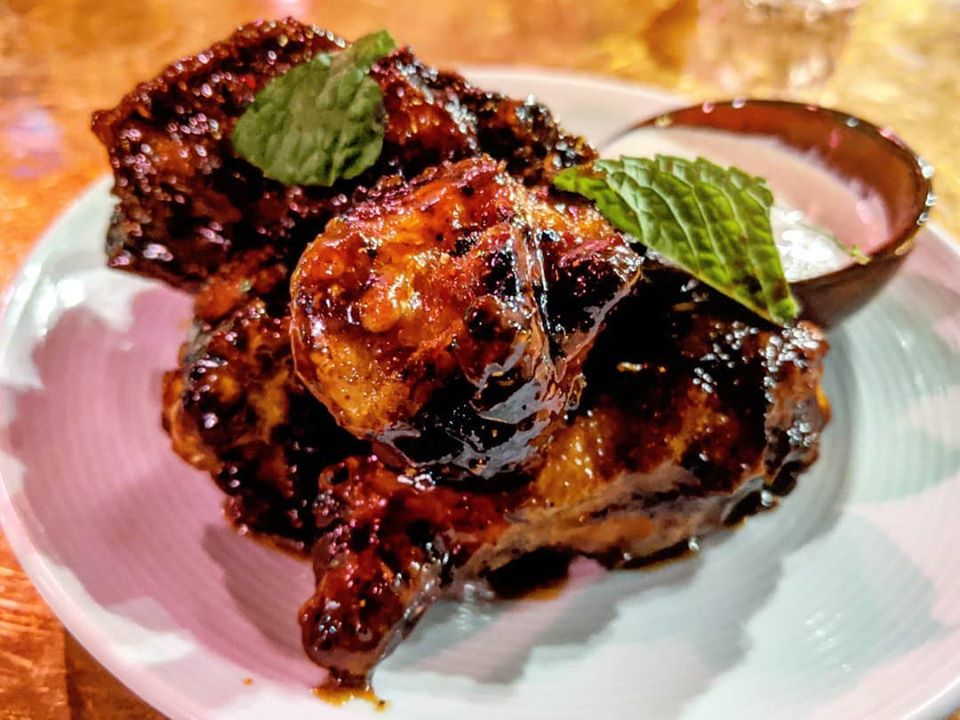 Sticky-looking, saucy, charred chicken wings sit on a plate, garnished with some mint leaves and served with a small dish of a dipping sauce.