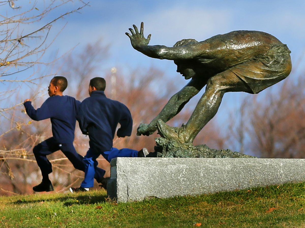 Bronze statue of a crouching man at the end of a long jump, and there’s two kids running by the statue.