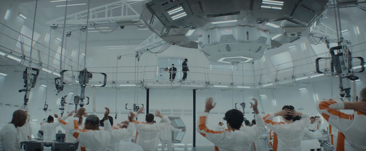 Prisoners in orange and white jumpsuits old their hands in the air below giant machinery as a new prisoner is escorted to the work area floor by way of a catwalk elevator in Andor
