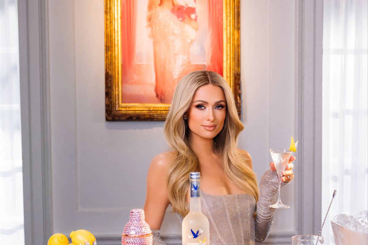 Paris Hilton stands behind a wooden bar with a bottle of Grey Goose, a pink cocktail shaker, and a blue bowl full of lemons. She is holding a martini.
